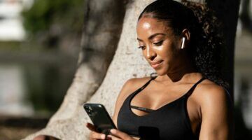 Vrouw met AirPods in die op haar telefoon zit terwijl ze tegen een boom aanleunt.