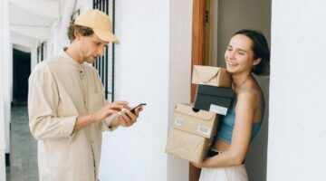 Een vrouw die veel pakketjes heeft ontvangen, en ze lachend vasthoudt. De postbode staat tegenover haar kijkend naar zijn telefoon.